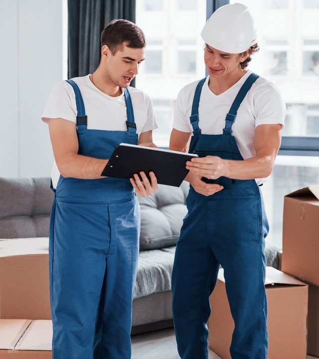 Holds notepad with document. Two young movers in blue uniform working indoors in the room.